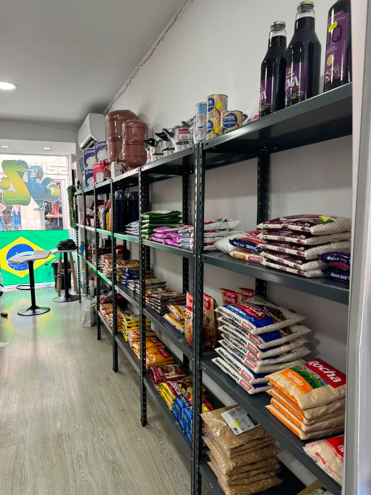 Prateleiras com farinhas, pacotes de grãos e bebidas, e o mural com a bandeira do Brasil ao fundo