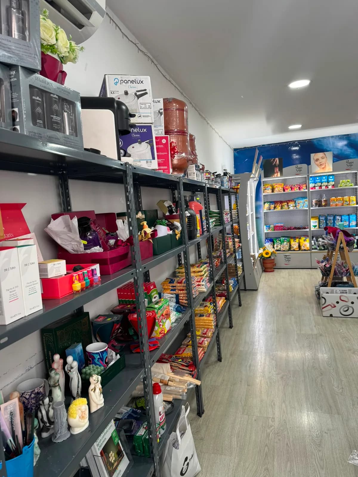 Corredor central da loja, com prateleiras de produtos brasileiros dos dois lados e a montra ao fundo