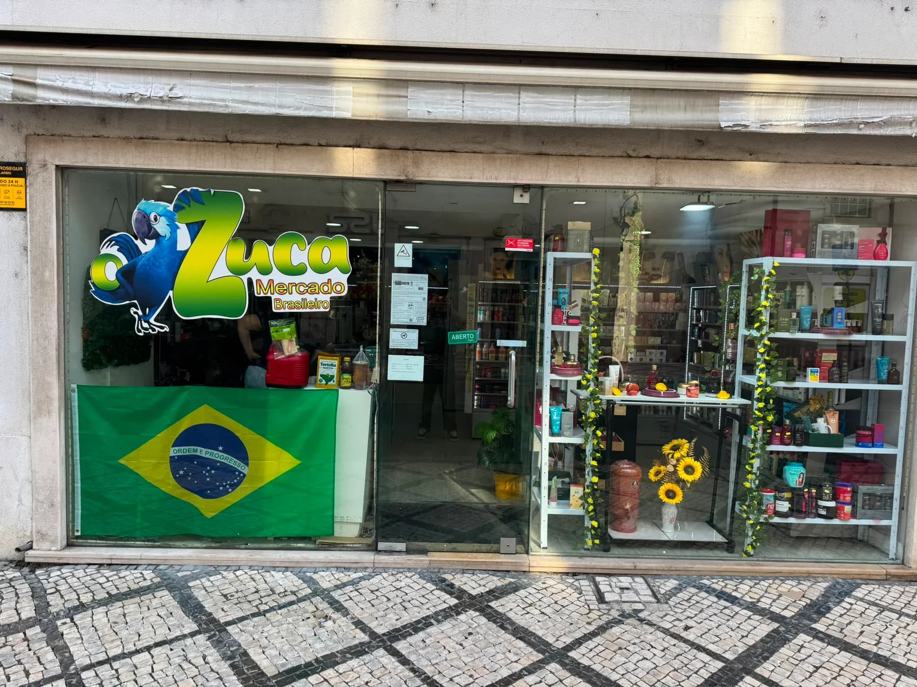 Fachada do Zuca Mercado Brasileiro na Rua da Sofia, em Coimbra, com o letreiro da arara azul, a bandeira do Brasil na montra e a loja aberta